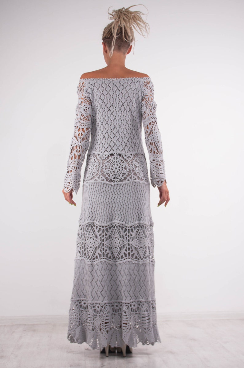 May include: A gray crochet maxi dress with a long-sleeved off-the-shoulder design. The dress features a delicate floral pattern and a tiered skirt.
