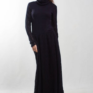 K&ouml;nnte beinhalten: Eine Frau tr&auml;gt ein langes, schwarzes Strickkleid mit Rollkragen. Das Kleid hat einen figurbetonten Oberteil und einen flie&szlig;enden Rock. Sie tr&auml;gt schwarze Schuhe mit Absatz.