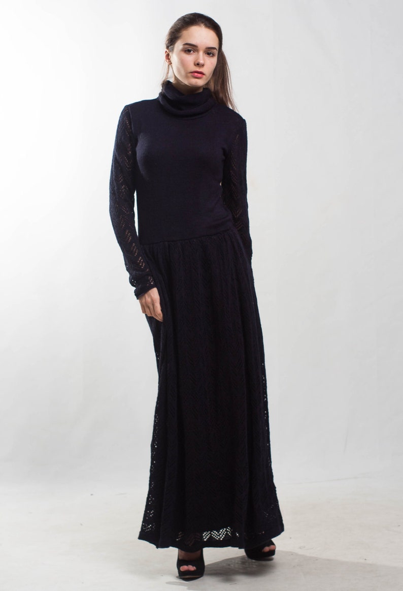 K&ouml;nnte beinhalten: Eine Frau tr&auml;gt ein langes, schwarzes Strickkleid mit Rollkragen. Das Kleid hat einen figurbetonten Oberteil und einen flie&szlig;enden Rock. Sie tr&auml;gt schwarze Schuhe mit Absatz.