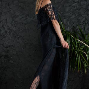 May include: A woman wearing a black crochet dress with a long, flowing skirt and a fringe detail. She is wearing black high-heeled sandals and has a tattoo on her leg.