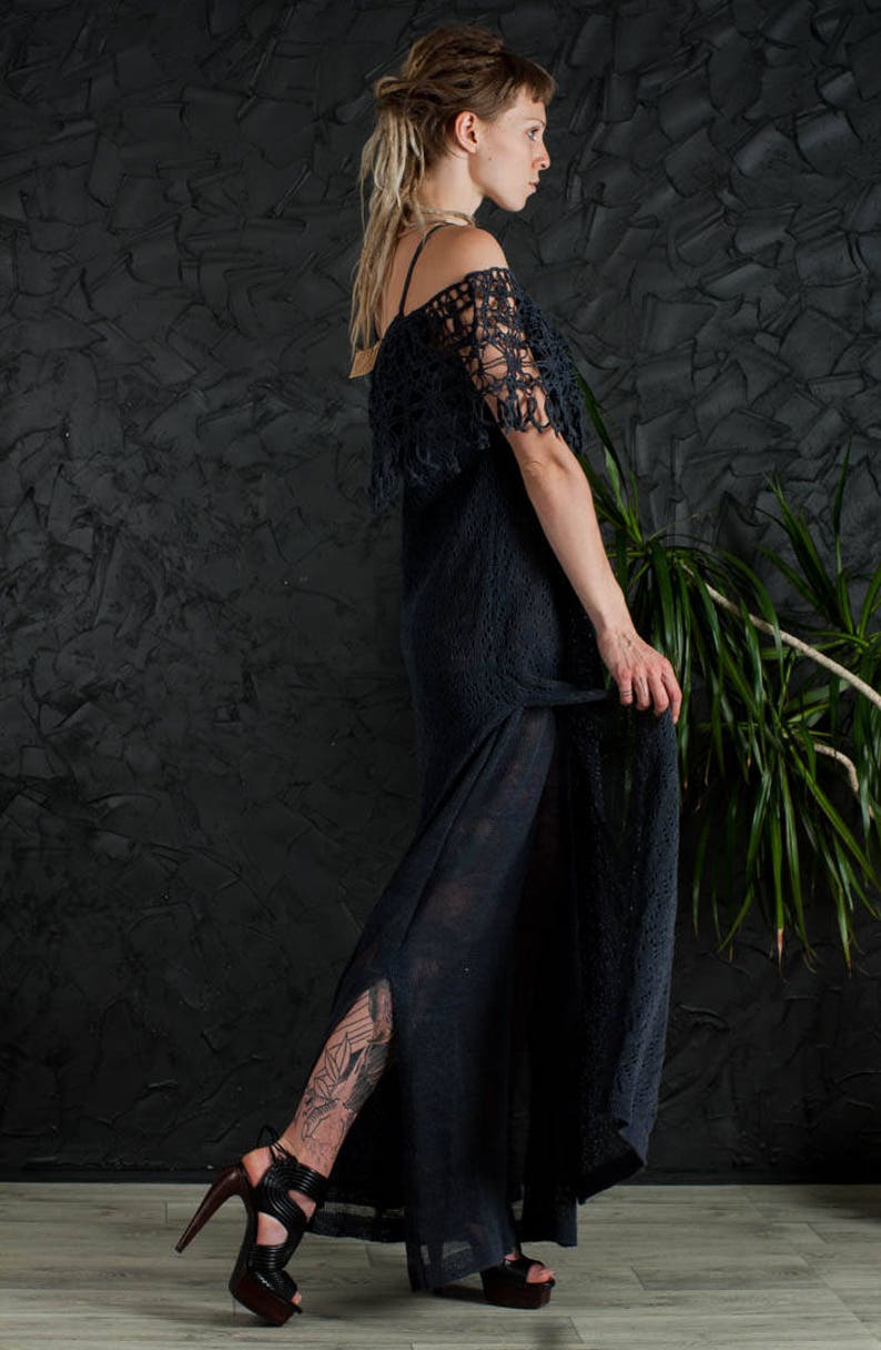 May include: A woman wearing a black crochet dress with a long, flowing skirt and a fringe detail. She is wearing black high-heeled sandals and has a tattoo on her leg.