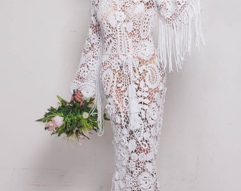 May include: A white, long crochet dress with a V-neck, long sleeves with fringe, and a floral pattern. The dress has a long train and is worn with a bouquet of pink and green flowers.