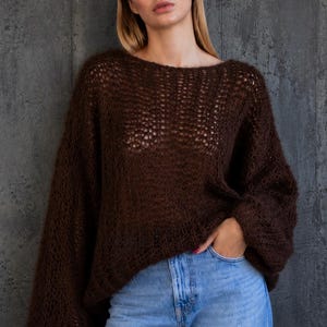 May include: A dark brown, loose-fitting, long-sleeved sweater with a wide boat neckline and a semi-sheer, open-knit texture. The garment features dropped shoulders and a relaxed silhouette, paired with light blue denim jeans.