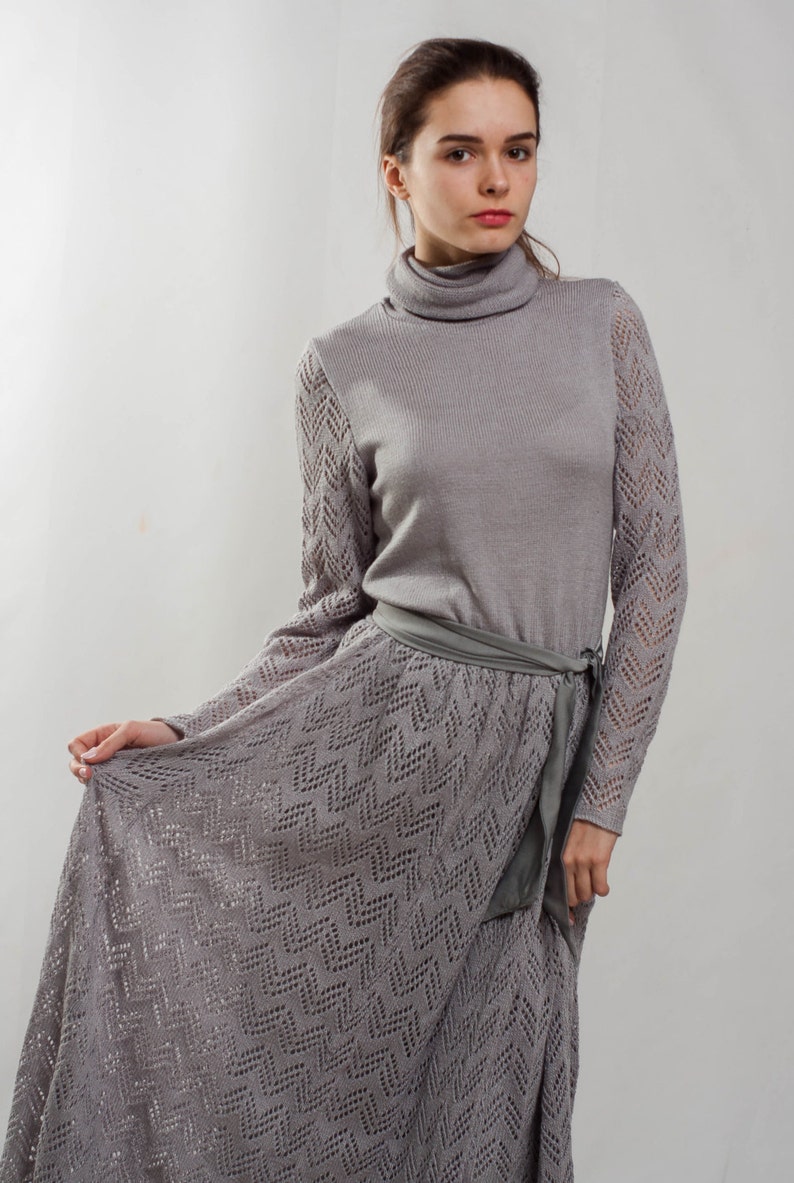 May include: A gray knitted dress with a long skirt and a turtleneck. The dress has a delicate lace pattern and a matching gray belt.
