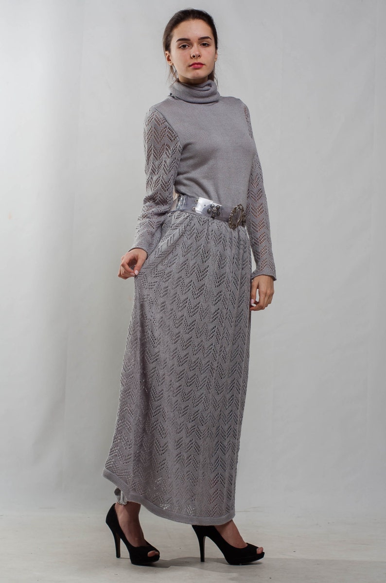 May include: A gray knitted maxi dress with a turtleneck and a silver belt. The dress has a chevron pattern and is worn by a person with dark hair.