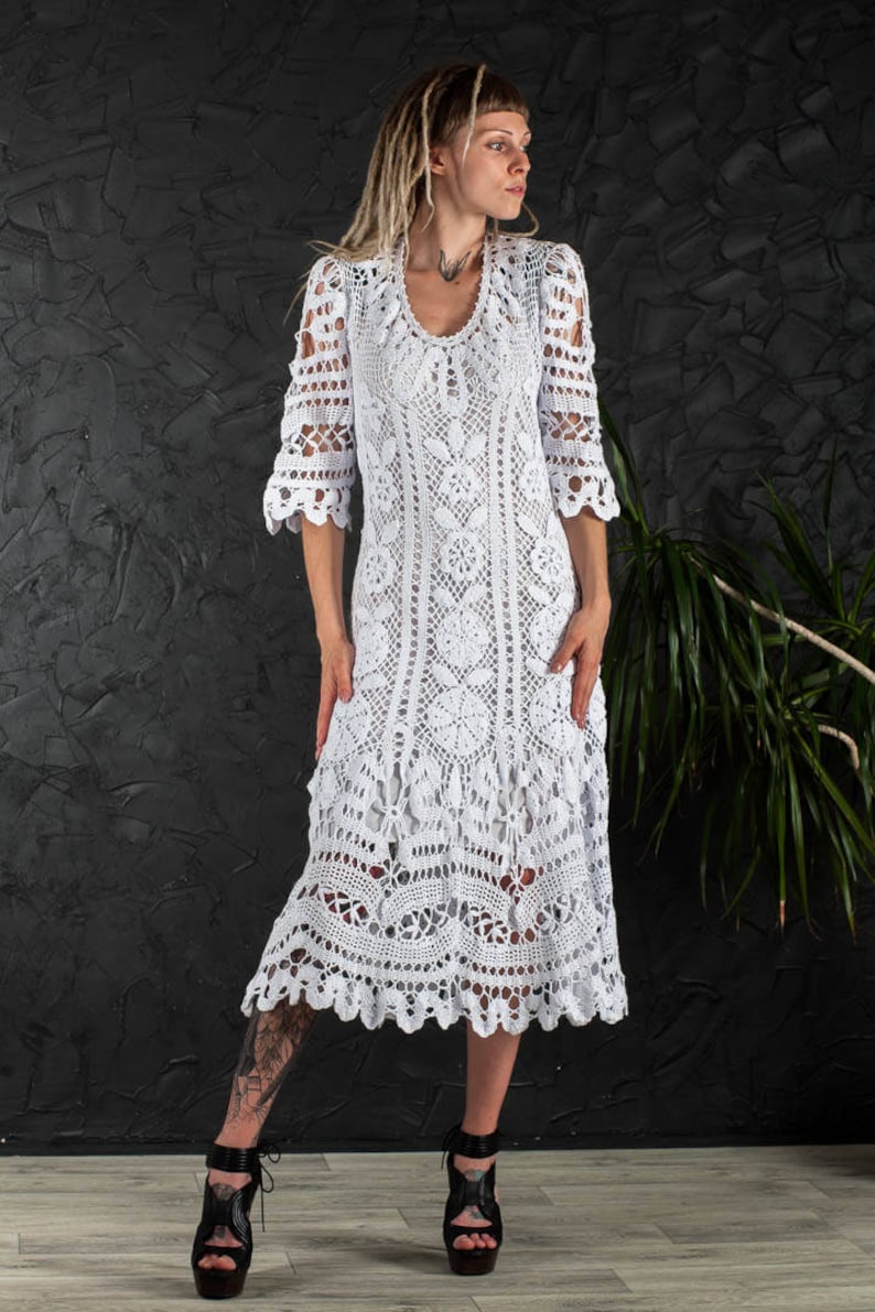 May include: A white crochet maxi dress with a delicate floral pattern. The dress has a scoop neckline and 3/4 sleeves. The dress is shown on a model with dreadlocks.