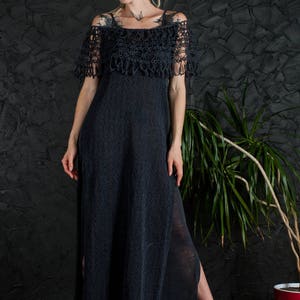 May include: A black crochet maxi dress with a lace overlay and a side slit. The dress is worn by a person with short hair and tattoos.