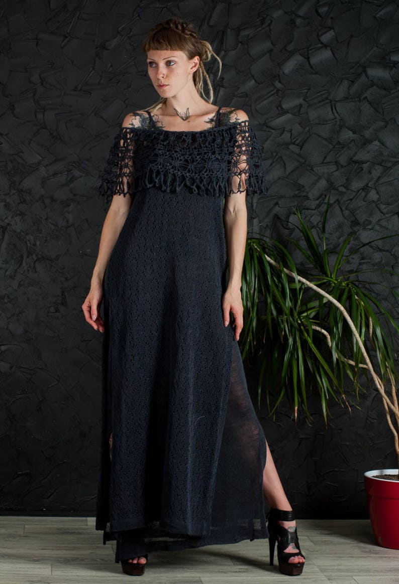 May include: A black crochet maxi dress with a lace overlay and a side slit. The dress is worn by a person with short hair and tattoos.