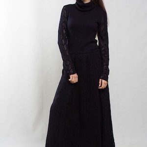 K&ouml;nnte beinhalten: Eine Frau tr&auml;gt ein langes, schwarzes Strickkleid mit hohem Kragen und langen &Auml;rmeln. Das Kleid hat ein spitzem Muster an den &Auml;rmeln und am Saum. Sie tr&auml;gt schwarze Abs&auml;tze.
