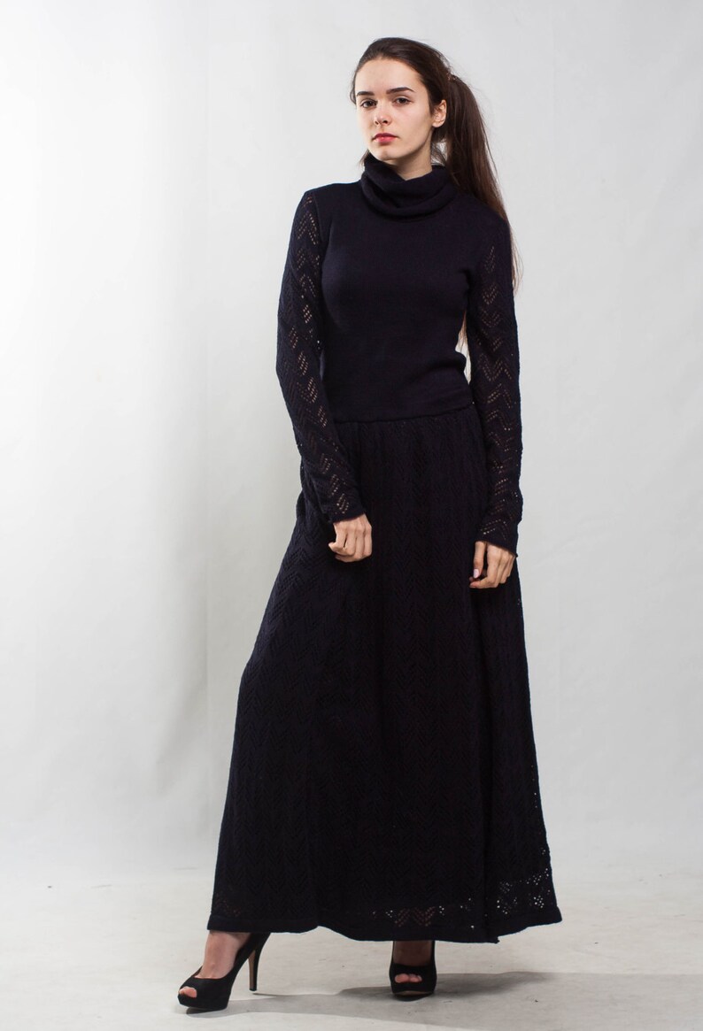 K&ouml;nnte beinhalten: Eine Frau tr&auml;gt ein langes, schwarzes Strickkleid mit hohem Kragen und langen &Auml;rmeln. Das Kleid hat ein spitzem Muster an den &Auml;rmeln und am Saum. Sie tr&auml;gt schwarze Abs&auml;tze.