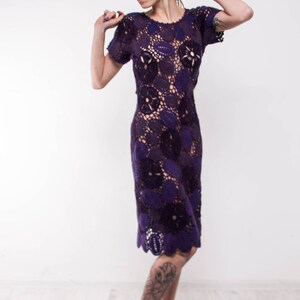 May include: A woman wearing a purple crochet dress with a floral pattern. She is wearing black high heels and has a tattoo on her leg.