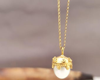 May include: A gold-colored necklace with a pendant shaped like an acorn. The pendant has a clear, round, pearl-like orb encased in a decorative gold cap. The chain is also gold and hangs against a blurred background.