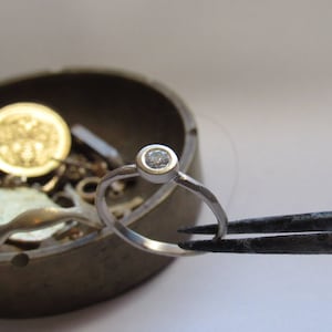 May include: A silver ring with a single, clear, round gemstone set in the centre. The ring is being held by a pair of tweezers.