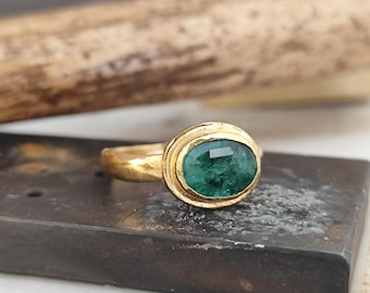 May include: A gold ring featuring an oval-cut emerald gemstone. The emerald is set in a bezel setting, and the ring band has a textured finish. The ring is displayed on a dark, textured surface, showcasing the jewelry's craftsmanship and design.