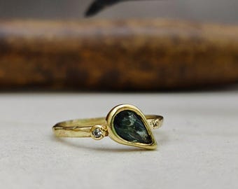 May include: A gold ring featuring a teardrop-shaped, dark green gemstone. A small, round, clear stone is set next to the main stone. The ring has a delicate band with subtle detailing.