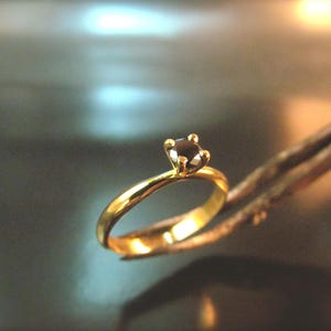 May include: A gold ring featuring a dark gemstone held by tweezers. The ring has a slender, polished band. The gemstone is set in a four-prong setting. The background is blurred, highlighting the ring's details.