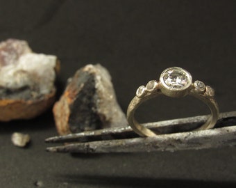 Könnte beinhalten: Ein silberner Ring mit einem großen runden Diamanten und drei kleineren Diamanten auf der Ringschiene.