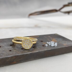 May include: A gold ring with a round, pave-set diamond centre stone. The ring is on a metal surface with loose diamonds scattered nearby.