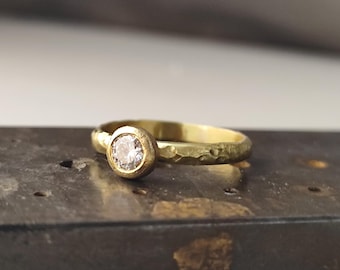 May include: A gold ring featuring a single round, clear gemstone set in a bezel setting. The band of the ring has a textured, hammered finish, giving it an uneven, organic appearance. The metal has a warm, yellow-gold tone throughout.