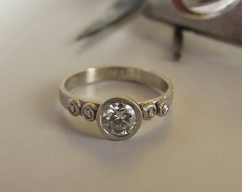 Könnte beinhalten: Ein silberner Ring mit einem großen runden Diamanten und kleineren Diamanten auf der Ringschiene.
