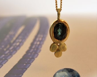 May include: A gold pendant necklace with a dark teal oval gemstone. The pendant has a gold setting and three small gold accents below the gemstone. Several loose gemstones in shades of blue and green are scattered below the pendant.