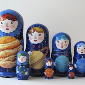 May include: A set of seven nesting dolls, each painted with a different astronaut wearing a blue spacesuit and holding a planet. The largest doll is holding Jupiter, the next is holding Saturn, the next is holding Neptune, and so on. The dolls are all painted in a whimsical style with bright colors.