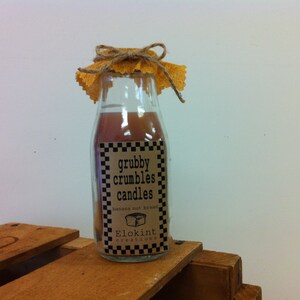 Könnte beinhalten: Ein Glasgefäß mit einer braunen Flüssigkeit darin. Das Gefäß hat ein braunes Etikett mit weißem Text, auf dem "grubby crumbles candles banana nut bread Elokin creations" steht. Das Gefäß ist mit Schnur und einem gelben Stoff gebunden.