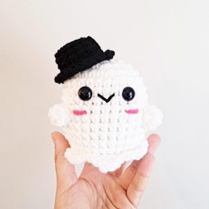 May include: A white crocheted ghost wearing a black top hat. The ghost has black eyes and pink cheeks.