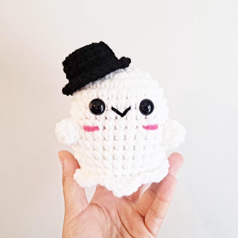 May include: A white crocheted ghost wearing a black top hat. The ghost has black eyes and pink cheeks.