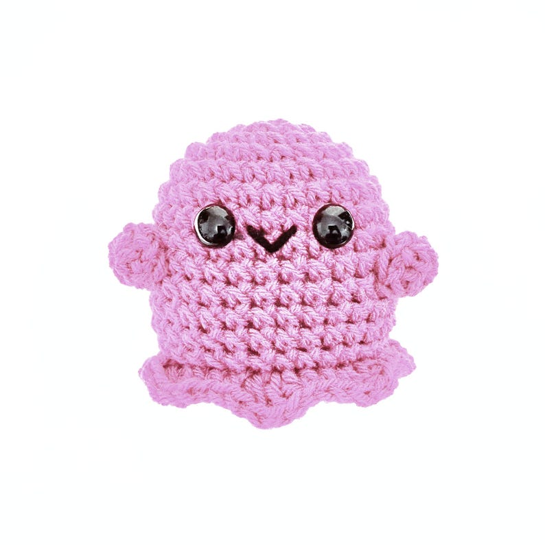 May include: A pink crocheted plush toy with a smiling face and black eyes. The toy is shaped like a blob or a small creature.