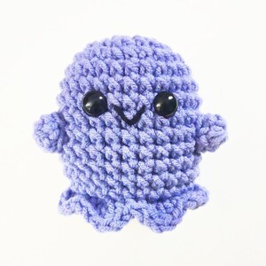 May include: A small, crocheted purple ghost with black eyes and a smiling mouth. The ghost has a rounded body and small arms and legs.