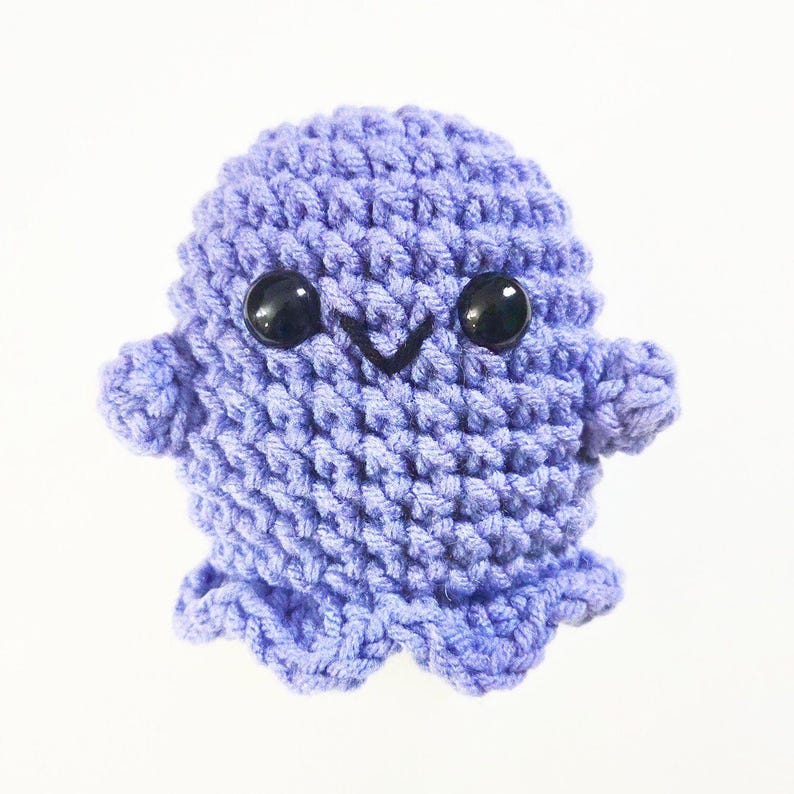 May include: A small, crocheted purple ghost with black eyes and a smiling mouth. The ghost has a rounded body and small arms and legs.
