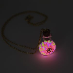 May include: A glowing glass pendant necklace with a gold chain. The pendant is a small, round bottle filled with colorful, glowing stones. A sun symbol is on the front of the bottle. The bottle is lit with a pink glow.