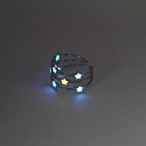 May include: A silver-toned metal ring featuring a multi-band design adorned with several star-shaped cutouts. The star cutouts emit a glowing light, with most stars appearing in a cool blue hue and one central star glowing in a warm yellow colour.