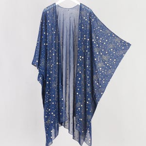 Pode incluir: Um quimono transparente azul-marinho com um padrão repetitivo de pequenas estrelas douradas. A peça tem a frente aberta, mangas largas e fluidas, e é feita de tecido leve e semitransparente pendurado num cabide de plástico branco.