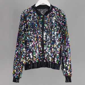 May include: A black sequin bomber jacket with a zipper closure, black trim, and long sleeves. The jacket is covered in colorful sequins, creating a shimmering effect. The jacket is hanging on a white hanger.