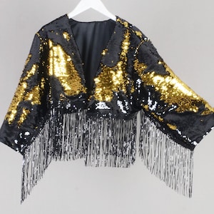 May include: A black sequin bolero jacket with gold accents and long silver fringe. The V-neck jacket is hanging on a white hanger. The garment is a party or special occasion top.