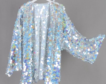 May include: A light blue sequin jacket with long sleeves and an open front. The jacket is covered in iridescent sequins that catch the light, creating a shimmering effect. The garment is hanging on a white hanger against a gray background.