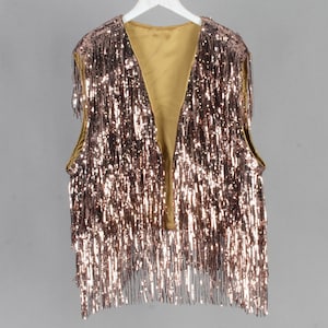 Puede incluir: Un chaleco sin mangas con cuello en V, cubierto de lentejuelas y flecos de color cobre brillante. El chaleco tiene un forro dorado y está colgado de una percha blanca. La prenda es adecuada para fiestas y eventos especiales.