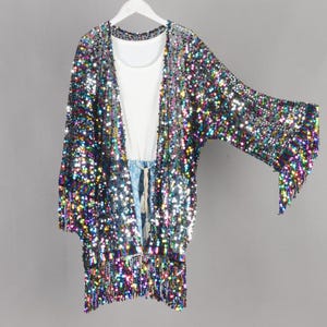 May include: A sequined kimono-style jacket with a white top underneath, hanging on a white hanger. The jacket is covered in iridescent sequins, with fringe at the hem and sleeves. The outfit is displayed against a gray background.