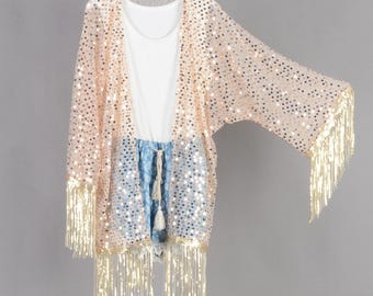 May include: A peach-coloured, sequined kimono with gold fringe, worn over a white top and blue shorts. The kimono has an open front. The shorts have a drawstring. The outfit is hanging on a white hanger.