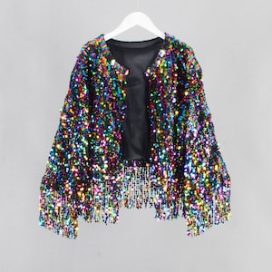 Puede incluir: Una chaqueta de lentejuelas arcoíris con ribete de flecos. La chaqueta está abierta en la parte delantera y tiene un forro negro.