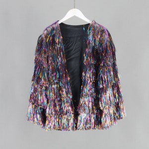 Könnte beinhalten: Mehrfarbige Jacke mit offenem Schnitt und mehrlagigen Fransen. Die Jacke hängt an einem weißen Kleiderbügel vor grauem Hintergrund. Die Fransen haben einen metallischen Glanz mit Farben wie Lila, Gold und Blau.