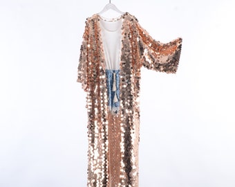May include: A long, rose gold sequined cardigan with a white top and denim shorts underneath. The cardigan is hanging on a white hanger against a white background.