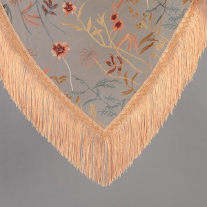 May include: A sheer, light gray scarf with embroidered floral patterns in shades of orange, yellow, and blue. The scarf has a peach-colored fringe trim along the edges, creating a delicate and elegant look. The floral design adds a touch of nature-inspired beauty.