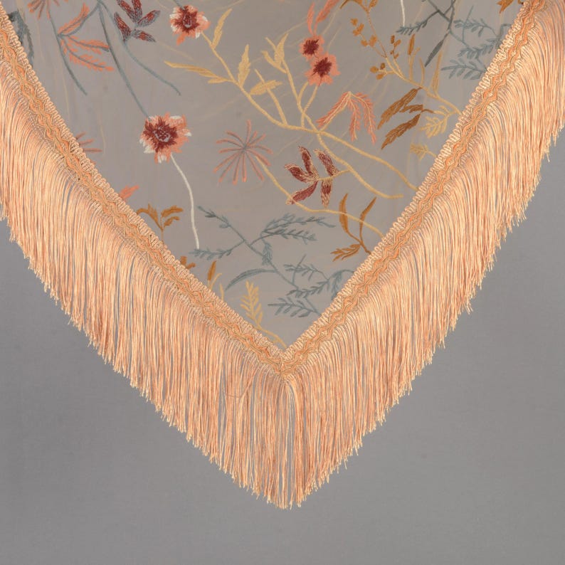 May include: A sheer, light gray scarf with embroidered floral patterns in shades of orange, yellow, and blue. The scarf has a peach-colored fringe trim along the edges, creating a delicate and elegant look. The floral design adds a touch of nature-inspired beauty.