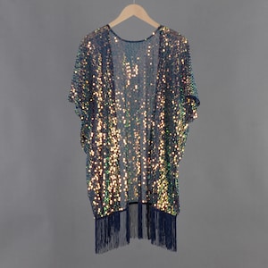 May include: A shimmering, sequined kimono in shades of teal and gold hangs on a wooden hanger. The open-front garment features short sleeves and a fringe trim along the bottom hem. The sequins create a sparkling effect, perfect for layering over an outfit.