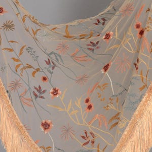 May include: A sheer, light gray scarf with embroidered floral patterns in shades of orange, peach, and yellow. The scarf has a delicate fringe trim in a matching peach color, adding a touch of elegance to the design.