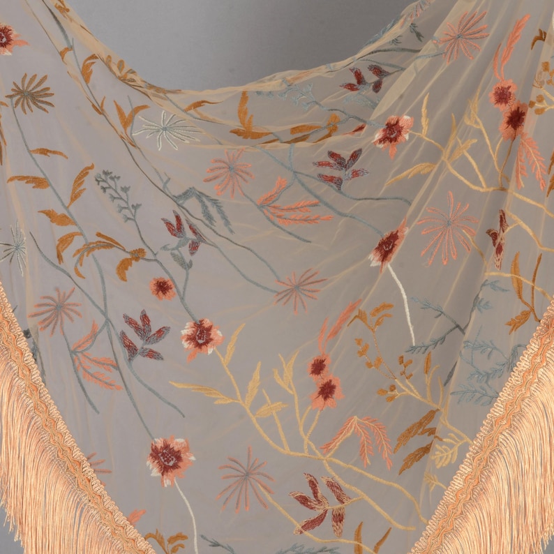 May include: A sheer, light gray scarf with embroidered floral patterns in shades of orange, peach, and yellow. The scarf has a delicate fringe trim in a matching peach color, adding a touch of elegance to the design.