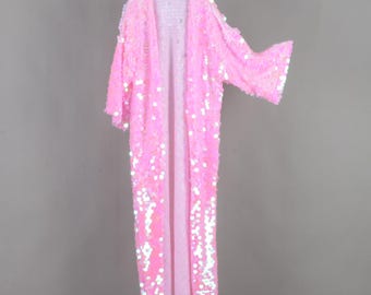 May include: A long, open-front pink sequin robe hanging on a white hanger. The robe is covered in small, shiny sequins, creating a shimmering effect. The sleeves are wide and flowing, and the robe is floor-length.